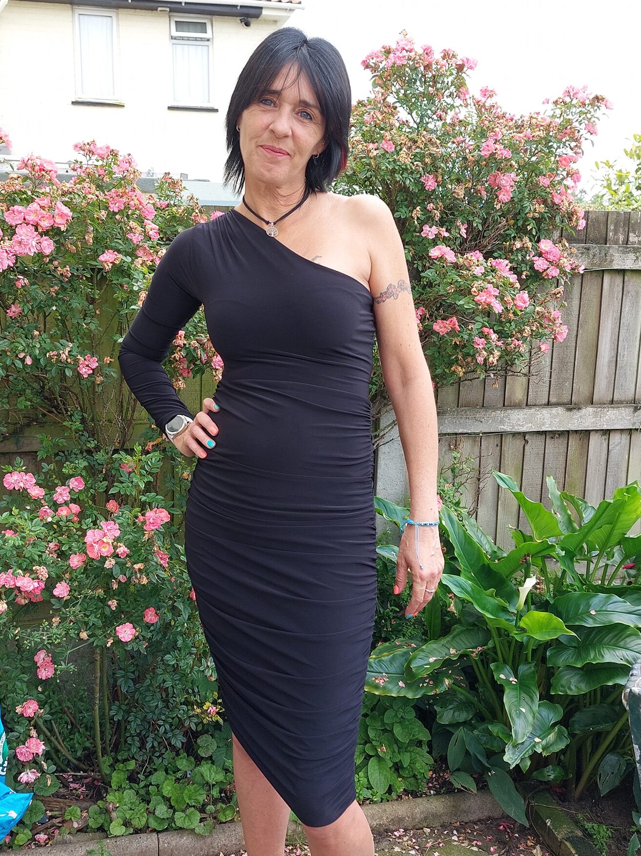 Lady wearing Black One Shoulder Ruched Slinky Dress in front of a shrub in the garden. The dress has one shoulder on it.