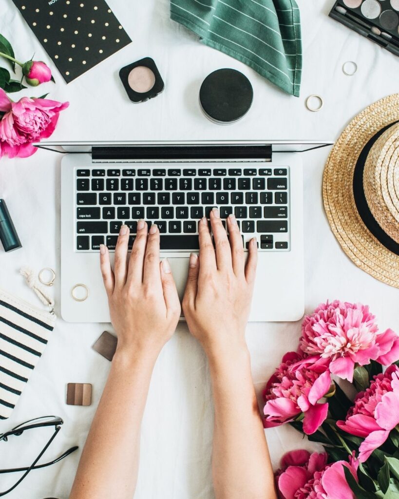A lady writing on her laptop, surrounded by flowers and a straw hat.