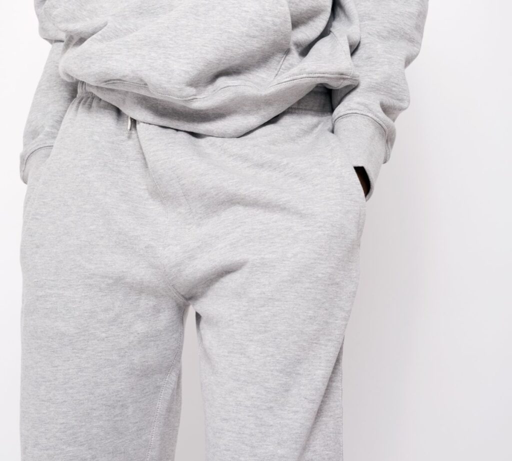 a man wearing a light grey jogging trousers and jumper combo. He is leaning against a white wall.